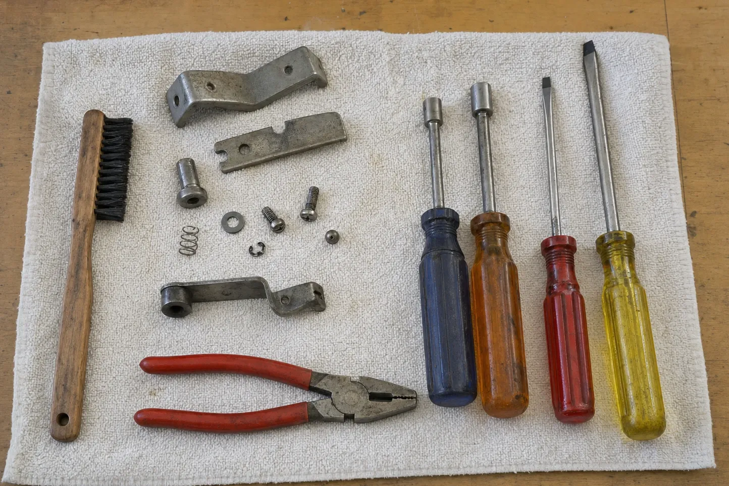 Oven door hinge arms, a spring, screws and washers laid out on a towel beside pliers, nut drivers and a wire brush